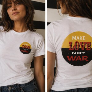 May include: A white short-sleeve t-shirt featuring a circular graphic on the front left chest and a larger version on the back. The graphic is split horizontally with yellow on top and black on the bottom, displaying the text MAKE LOVE NOT WAR.