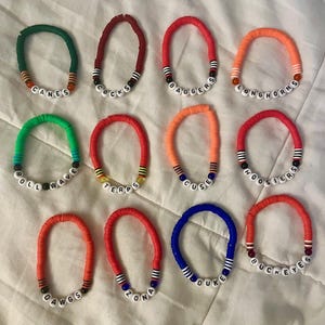 Puede incluir: Una colección de pulseras de cuentas de colores. Cada pulsera presenta un nombre de equipo diferente escrito con cuentas de letras blancas. Las pulseras son de varios colores, incluyendo rojo, verde, naranja y azul, con acentos decorativos.