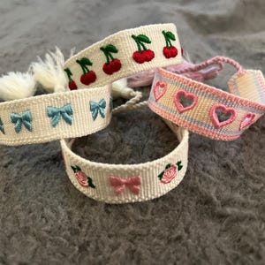 May include: Four woven friendship bracelets with different designs. One bracelet features pink hearts, another has blue bows, another has cherries, and the last one has pink bows and roses.