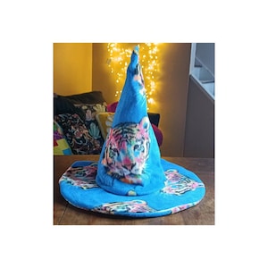 May include: A blue witch hat with a colorful tiger face design. The hat has a wide brim and a pointed top. The tiger's face is depicted with vibrant pink, yellow, and blue hues. The brim also features a repeating tiger face pattern.
