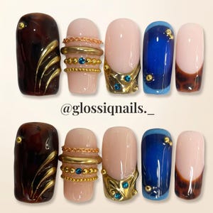 Puede incluir: Un juego de diez uñas postizas con diseños variados que incluyen carey marrón oscuro con detalles dorados, bases nude con cadenas y cuentas metálicas, adornos azules y dorados, y puntas azules. El texto @glossiqnails._ es visible.