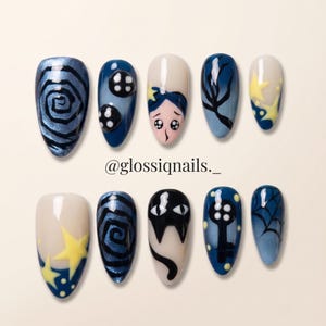 May include: A set of ten almond-shaped press-on nails featuring a dark blue, black, and cream color palette with hand-painted designs including spirals, buttons, a character face, stars, a cat face, a key, and a spider web. Text reads @glossiqnails._