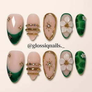 Puede incluir: Un juego de diez uñas postizas con base nude y puntas verde oscuro, detalles metálicos dorados, pequeños brillantes y flores blancas en relieve. El texto @glossiqnails._ aparece centrado entre las dos filas de uñas postizas.