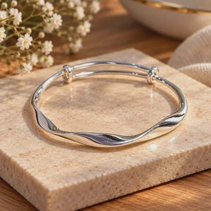 Könnte beinhalten: Ein silberfarbener Armreif mit einem glatten, wellenförmigen Design. Das Armband ist mit einem verstellbaren Schiebemechanismus ausgestattet, der durch zwei kleine kreisförmige Knoten gekennzeichnet ist, wodurch sich das Band weiten und verengen lässt.