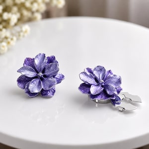 May include: A pair of purple flower-shaped earrings with a textured, metallic finish. One earring is shown from the front, while the other is positioned to reveal the silver-toned metal clip-on mechanism attached to the back of the floral design.