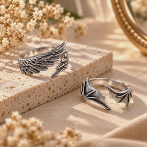 Puede incluir: Dos anillos de metal en tono plateado. Un anillo presenta un diseño detallado de alas de ave con plumas que envuelven el dedo. El segundo anillo presenta un diseño de alas de murciélago con puntas afiladas y membranas bien definidas.