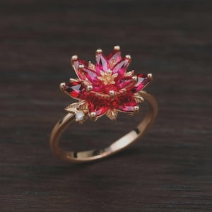 May include: A gold ring featuring a large, multi-layered flower design composed of marquise-cut red gemstones. The center of the flower has small gold stamens, and a single small, clear round gemstone is set on the band near the flower base.