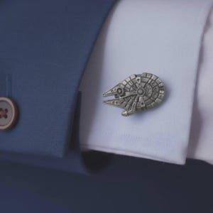 Könnte beinhalten: Ein silberfarbener Manschettenknopf in Form eines kreisförmigen Raumschiffs mit komplexen Paneelen und einer spitz zulaufenden Vorderseite, befestigt an der weißen Stoffmanschette eines Hemdes neben einem dunkelblauen Blazerärmel mit braunem Knopf.