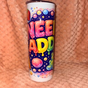 May include: A cylindrical metal tumbler featuring a vibrant, multi-coloured graphic print against a dark blue background. The design includes the text "NEED ADD" in large, bubble-style letters, surrounded by colourful candy-like shapes, bubbles, and stars.