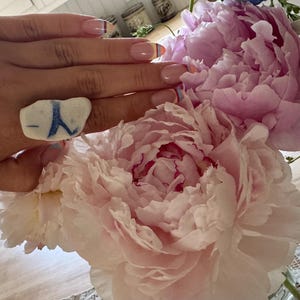 May include: A ring featuring a smooth, white, irregularly shaped piece of sea glass with a distinct blue letter Y marking on its surface. The ring is worn on a finger, positioned next to a large bouquet of light pink and white peony flowers.