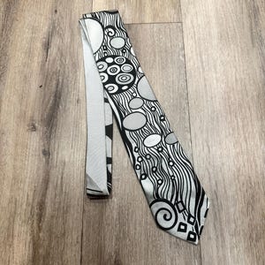 May include: A necktie featuring a black, white, and grey abstract pattern of swirling lines, circles, and geometric shapes. The fabric has a smooth finish, and the design covers the entire length of the tie, including the pointed end and the folded back.