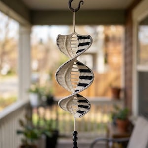 May include: A hanging crochet spiral ornament featuring a repeating DNA-like structure with alternating cream and black striped sections. The piece is suspended from a metal hook and finished at the bottom with two dark beads and a black tassel.