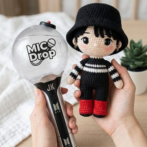 Puede incluir: Un bastón de luz plateado y negro con el texto MIC Drop y JK impreso, junto a una pequeña muñeca de ganchillo que lleva un sombrero de pescador negro, una camiseta de rayas negras y blancas, pantalones negros y zapatos rojos con aros pequeños.
