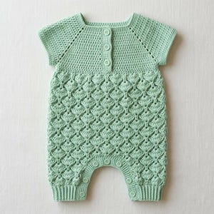 Puede incluir: Un pelele de ganchillo en color verde menta claro con un patrón de puntos de concha texturizados en el cuerpo y mangas cortas. La prenda tiene un cuello redondo con una tapeta vertical de tres botones pequeños a juego y una abertura abotonada.