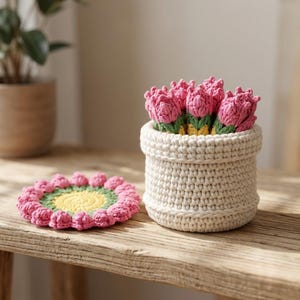 May include: A cream-colored crocheted basket filled with several pink crocheted tulip flowers with green stems, accompanied by a matching circular crocheted coaster featuring a yellow center, green ring, and pink petal-like border.