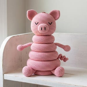May include: A pink crocheted pig stacking toy featuring a pig head with closed eyes and a snout, three circular rings of varying sizes, two small arms, and a curly tail. The toy is constructed from yarn with a visible textured stitch pattern.