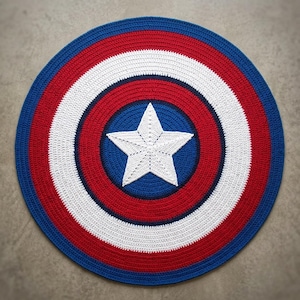 May include: A circular crochet rug featuring a concentric pattern of blue, red, and white rings. The center of the rug is a solid blue circle with a large, five-pointed white star stitched in the middle, creating a classic shield-like design.