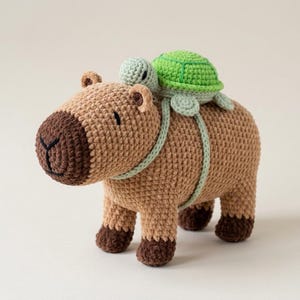 Pode incluir: Uma capivara de crochê bege com focinho e patas marrom-escuro, com uma pequena tartaruga de crochê verde e verde-claro nas costas. A tartaruga está presa com tiras de fio verde-claro que envolvem o corpo da capivara.