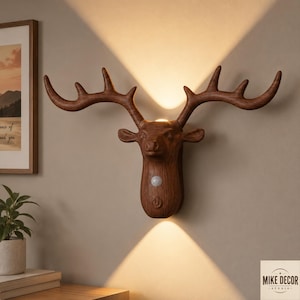 May include: A wall-mounted deer head lamp with a wood-grain finish, featuring a motion sensor and a power button on the front. The light fixture emits a warm glow directed both upwards and downwards, creating two distinct triangular light beams.