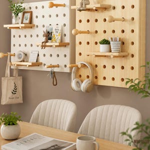 May include: Two rectangular pegboard wall organisers, one white and one light wood, featuring adjustable wooden shelves and pegs. The boards hold small potted plants, a framed picture, a book titled Every Day, a clock, keys, and headphones.