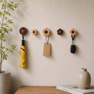 May include: Five wall-mounted wooden hooks in various shapes and sizes, including circular and flower-shaped designs. The hooks feature a two-tone wood finish with dark brown centres and lighter wood edges, holding items like an umbrella and keys.