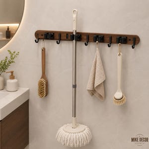 May include: A wall-mounted storage rack featuring a dark wood grain finish with multiple black hooks and spring-loaded clips. The rack holds a mop with a white cotton head, a wooden scrub brush, a beige cloth, and a long-handled cleaning brush.