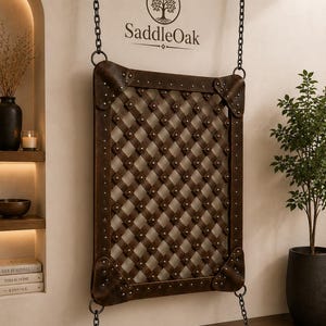Puede incluir: Un expositor de pared rectangular con un panel de celosía trenzado de color marrón oscuro y tachuelas metálicas. El panel está suspendido por cadenas de metal negro de una barra horizontal superior y anclado a una barra inferior, con el texto SaddleOak.