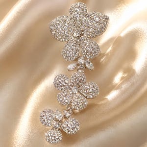May include: A silver-toned brooch featuring three connected flower shapes, each densely encrusted with clear, sparkling crystals. The design includes small marquise-cut stones between the floral elements, creating a cascading, vertical arrangement.