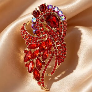 May include: A gold-tone metal brooch featuring a large teardrop-shaped red crystal at the top, surrounded by numerous smaller round and marquise-cut red crystals. The design includes accents of iridescent, multi-coloured round crystals.