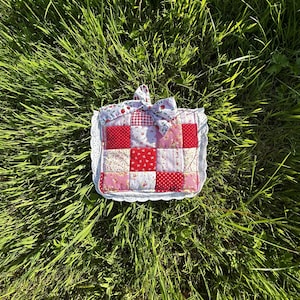 May include: A square quilted potholder featuring a patchwork design of red, pink, and white fabrics with floral and gingham patterns. The potholder is finished with a white eyelet lace trim and a matching fabric bow attached to the top edge.