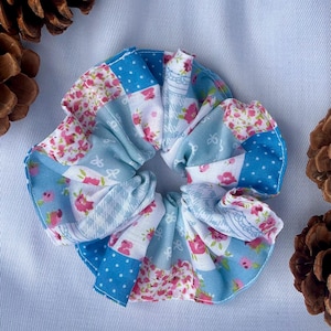Può includere: Un elastico per capelli in tessuto con un design patchwork caratterizzato da pannelli alternati con motivi floreali rosa, bianchi e azzurri, pois blu e sottili stampe geometriche bianche, raccolto in una forma circolare arricciata.