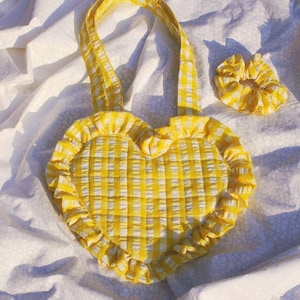 May include: A heart-shaped quilted tote bag and a matching scrunchie, both featuring a yellow and white gingham check pattern. The tote bag has a ruffled border along its edge and two fabric handles made from the same yellow and white fabric.