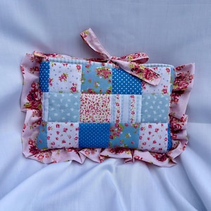 Può includere: Un cuscino patchwork rettangolare caratterizzato da una griglia di quadrati in tessuto blu e rosa con motivi floreali, a pois e a righe. Il cuscino è incorniciato da un bordo arricciato floreale rosa e include un laccio in tessuto abbinato.