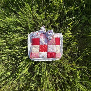 May include: A square quilted potholder featuring a patchwork design of red, pink, and white fabrics with floral and gingham patterns. The potholder is finished with a white eyelet lace trim and a matching fabric bow attached to the top edge.