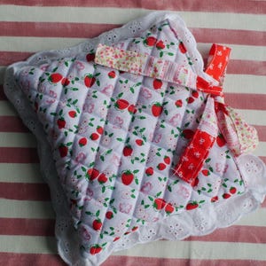 Könnte beinhalten: Ein quadratischer, gesteppter Topflappen mit weißem Stoffgrund und einem wiederkehrenden Muster aus roten Erdbeeren, grünen Blättern und kleinen rosa Schmetterlingen. Die Ränder sind mit weißer Lochstickerei und einer roten Aufhängeschlaufe versehen.