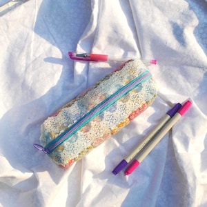 Op de afbeelding: Een rechthoekig etui van stof met een veelkleurig bloemenpatroon, een witte kanten overlay en een felblauwe rits. Drie pennen met roze en paarse accenten liggen rondom het etui op een ondergrond van witte stof.