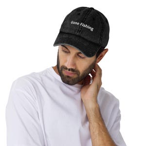 May include: A black baseball cap with a distressed, washed-out finish. The front of the cap features the white embroidered text "Gone Fishing" in a clean, sans-serif font. The cap has a curved brim and a structured crown with visible stitching.