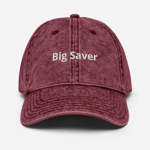 May include: A maroon baseball cap with a vintage washed finish and a structured six-panel design. The front features the text Big Saver embroidered in white thread. The cap includes a curved brim and a button at the top center of the crown.
