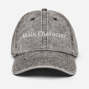 May include: A charcoal grey acid-wash baseball cap with a curved brim and six-panel construction. The front features the text Main Character embroidered in white thread. The cap includes a top button and stitched eyelets for ventilation.