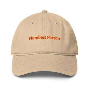 May include: A beige baseball cap with a structured crown and a curved brim. The front of the cap features the text "Numbers Person" embroidered in orange thread. The hat includes stitched eyelets on the crown and decorative stitching on the brim.