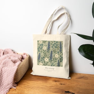 May include: A cream-colored canvas tote bag featuring a square botanical print of blue hyssop flowers, white blossoms, and green leaves on a sage green background. The text Hyssop, Cleansing, Renewal, Mercy, Psalm 51:7 is printed below the design.