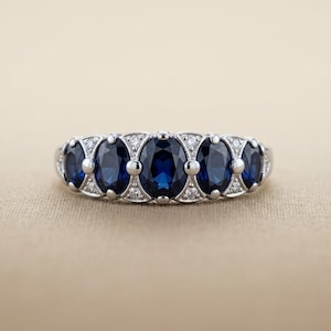 May include: A silver-toned ring featuring five oval-cut deep blue sapphires arranged in a row. The stones are separated by small, round silver beads and accented with tiny, clear, round-cut diamonds set within the metal framework between each sapphire.