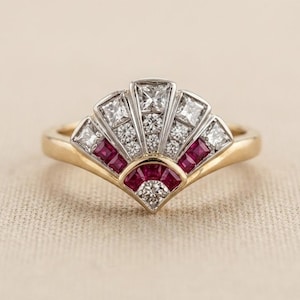 May include: A gold ring featuring a fan-shaped centerpiece set with multiple square-cut rubies and clear diamonds. The design includes a central row of round diamonds flanked by princess-cut diamonds, all arranged in a radiating, geometric pattern.
