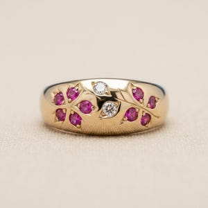 Op de afbeelding: Een gouden ring met een gepolijst oppervlak en gegraveerde bladvormige patronen. Het ontwerp is geaccentueerd met acht ronde, felroze robijnen en twee heldere, rond geslepen witte diamanten die in het metaal zijn gezet.