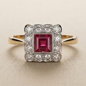 Op de afbeelding: Een gouden ring met een vierkant geslepen dieprode edelsteen in het midden, omringd door een geschulpte halo van kleine ronde witte steentjes in een zilverkleurige zetting. De ringband is van glad, gepolijst goud en verbonden met het midden.
