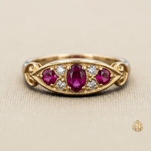 Könnte beinhalten: Ein Ring aus Gelbgold mit einem zentralen oval geschliffenen Rubin, flankiert von zwei kleineren runden Rubinen und vier kleinen runden Diamanten. Die Fassung hat einen dekorativen Milgrain-Rand und Schnörkel an den Seiten der Ringschiene.