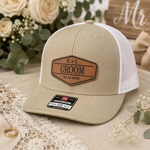 May include: A tan and white trucker hat featuring a brown leather patch on the front. The patch is hexagon-shaped and engraved with the text E + L, GROOM, and the date 06.14.2026. A black and white sticker is attached to the flat brim of the hat.