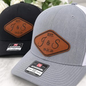 May include: Two adjustable trucker caps, one black and one grey with white mesh, each featuring a brown diamond-shaped patch. The patch is engraved with the text EST. J & S 09.21.26. A black sticker on the brim displays the Richardson 112 logo.