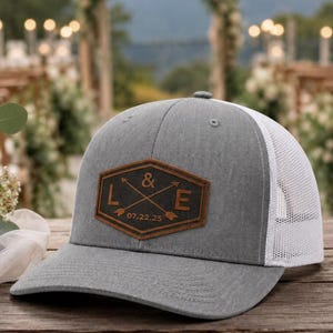 May include: A gray baseball cap with a white mesh back and a brown leather patch on the front. The patch is hexagonal and features the letters L and E separated by an ampersand and crossed arrows, with the date 07.22.25 engraved below.