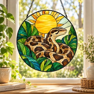 May include: A circular stained glass suncatcher featuring a coiled brown and tan snake surrounded by vibrant green tropical leaves. A bright yellow sun with radiating beams sits in the background. The piece is framed in black and hangs by a chain.